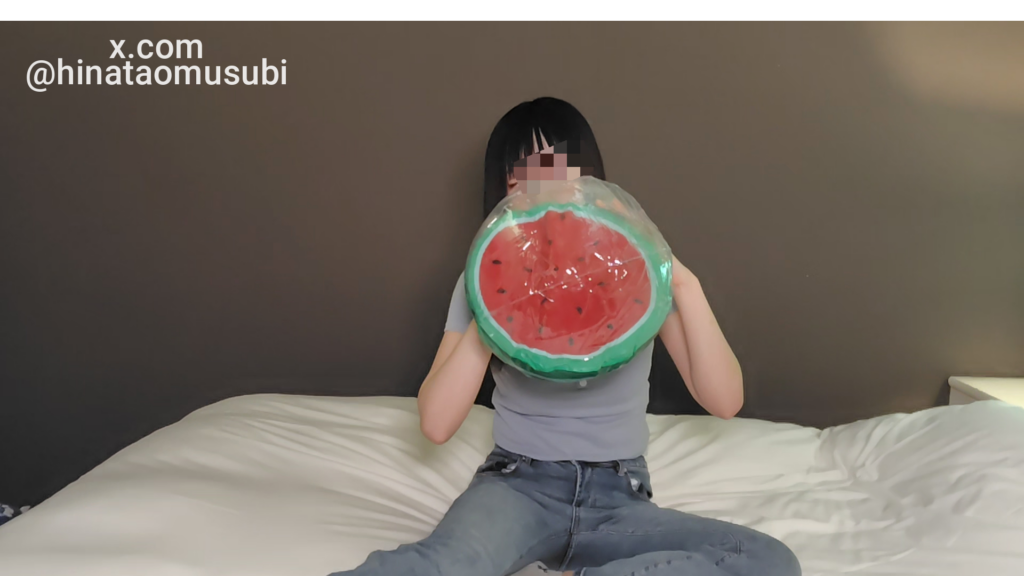 ベッドに座る女性が、大きく膨らましたスイカ柄インフレータブルボール(赤い果肉面)を顔の前で両手で抱えている