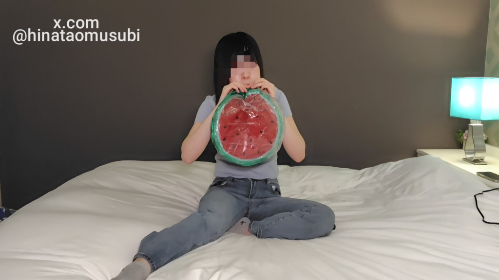 ベッドに座る女性が、スイカ柄ビーチボールの口を自分の口に近づけて空気を吹き込んでいる様子